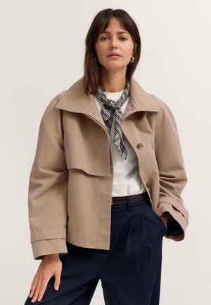 Frau trägt beige gekürzten Trenchcoat, weißes Oberteil, marineblaue Hose, gestreiften Halstuch und goldene Creolen, steht mit der Hand in der Tasche.