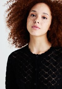 Cardigan tricoté noir avec un motif en losanges texturé, col rond et fermeture par boutons. Le tissu est léger et à mailles ouvertes.