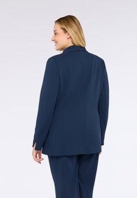 Kvinna med blont hår klädd i en mörkblå kavaj och matchande byxor, vänd lite åt sidan med en ljusgrå bakgrund.