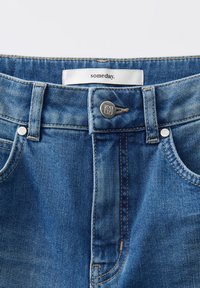Gros plan sur la taille d'un jean en denim bleu avec un bouton en métal, des passants de ceinture, des rivets et une étiquette blanche portant l'inscription "un jour."