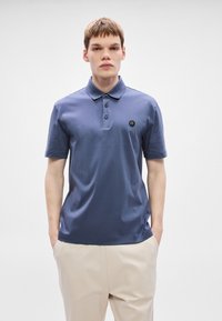 Námornícka polokošeľa s krátkymi rukávmi, zapínaním na tri gombíky a malým logom na ľavej strane hrudníka; v kombinácii s svetlo béžovými nohavicami.