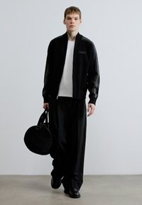 Zwarte jas met rits en grijze strepen, gecombineerd met een wit shirt en losse zwarte broek. Man draagt een ronde zwarte tas en zwarte schoenen.