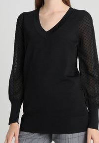 Pull-over noir à col en V, corps uni, manches bouffantes transparentes à texture ponctuée. Matière en tricot doux avec détails côtelés.