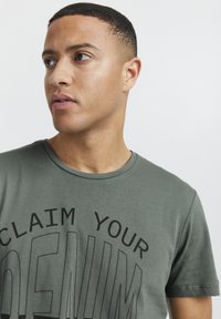 Grønn t-skjorte i bomull med rund hals og fet svart tekst som sier "CLAIM YOUR DENIM," med en jevn tekstur.
