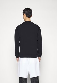 Černá mikina s dlouhými rukávy, kulatým výstřihem a žebrovanými manžetami, kombinována s bílými šortkami a pruhovanými kalhotami, viděná zezadu.