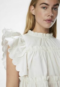 Abito bianco con maniche a volant scallopate, scollatura raccolta e collo alto. Realizzato in tessuto leggero con una trama liscia.