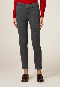 Femme portant un jean gris slim avec de petits clous en argent, un haut rouge à manches longues et des mocassins rouge foncé avec des franges.