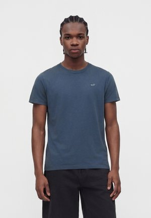 Junger Mann mit geflochtenem Haar, der ein schlichtes blaues T-Shirt und schwarze Hosen trägt, steht vor einem einfarbigen hellen Hintergrund.