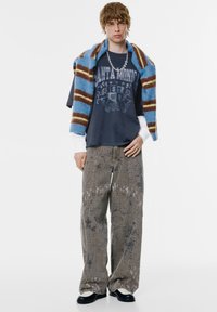 Szürke grafikás póló "Santa Monica" felirattal, laza mintás bézs nadrág, és kék-barna csíkos pulóver a vállon. Fekete cipők.