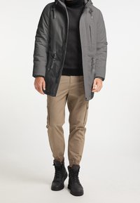 Veste isolante gris foncé avec une fermeture éclair à l'avant, une capuche et des cordons de serrage, portée par-dessus un pull noir et un pantalon cargo beige clair avec des poignets à la cheville.