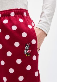 Κόκκινο παντελόνι από ύφασμα με μεγάλα λευκά πουά και κεντημένο καρτούν χαρακτήρα κοντά στην τσέπη, με το χέρι μερικώς μέσα στην τσέπη.