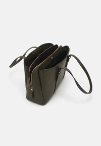Sac fourre-tout en cuir vert olive avec deux compartiments, fermeture éclair dorée, extérieur texturé et poignées doubles. Intérieur doublé avec une finition sombre.