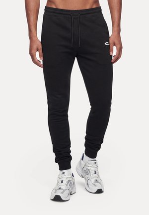 Männlicher Oberkörper und Beine in schwarzen Jogginghosen mit kleinem weißen Logo und weißen New Balance Sneakern vor schlichtem Hintergrund.