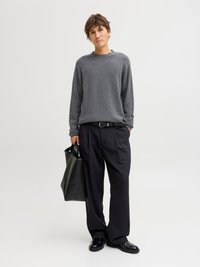 Joven con un suéter gris, pantalones negros sueltos, zapatos negros, sosteniendo un bolso tote negro, de pie frente a un fondo blanco liso.