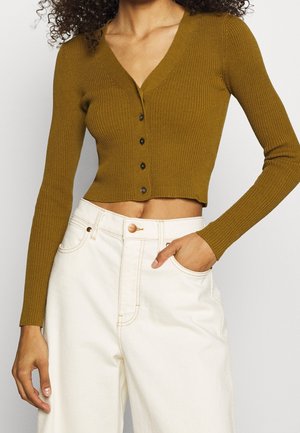 Cárdigan corto de canalé en verde oliva con mangas largas, escote en V y cinco botones oscuros en el frente, combinado con pantalones blancos de cintura alta.