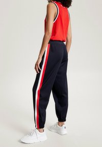 Marinträningsbyxor i marinblått med röda och vita sidostråk, elastisk midja och avsmalnande anklar, ihop med en röd ärmlös topp och vita sneakers.