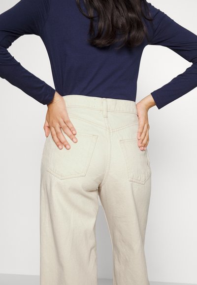 Beżowe spodnie o szerokich nogawkach z teksturowanej tkaniny, z tylnymi kieszeniami i wysokim stanem. W komplecie z granatową bluzką z długim rękawem.