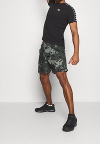 Černé tričko s krátkým rukávem a logem, doplněné o maskáčové šortky v tmavě zelené barvě. Model má na sobě černé a šedé sportovní boty.