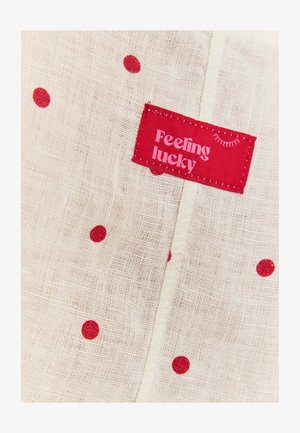 Bežs audums ar sarkaniem punktiem un sarkanu birku, uz kuras ir rakstīts "Jūtos laimīgs" rozā tekstā, ar vienkāršu šūšanas detaļu.