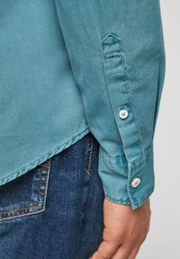 Chemise turquoise à manches longues en tissu à texture lisse ; elle présente des poignets boutonnés et un ourlet arrondi, associée à un jean en denim.