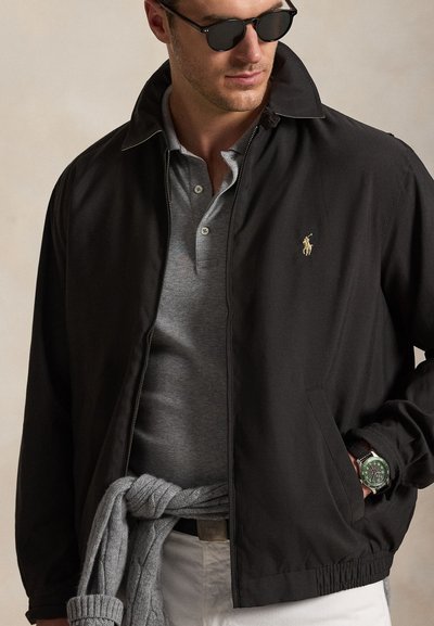 Férfi fekete dzsekit visel arany Polo logóval, szürke galléros pólót, megkötött szürke pulóvert, fehér nadrágot, napszemüveget és zöld számlapos karórát.