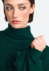 Grüner gerippter Strickpullover mit hohem Rollkragen. Der Stoff wirkt weich und strukturiert, mit langen Ärmeln und einer lässigen Passform.