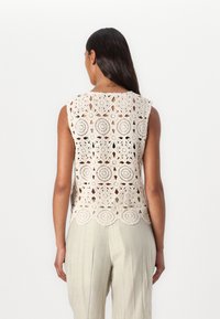 Hevnjakke i off-white med blomsterprint, viser åpen arbeidsdesign og bølgekant. Kombinert med lette, løstsittende bukser.