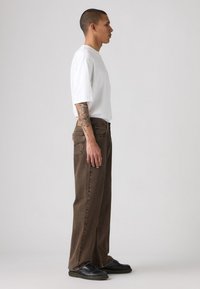 Hombre con brazo tatuado que lleva una camiseta blanca de gran tamaño, pantalones vaqueros sueltos marrones y zapatos negros, de pie en perfil contra un fondo liso.