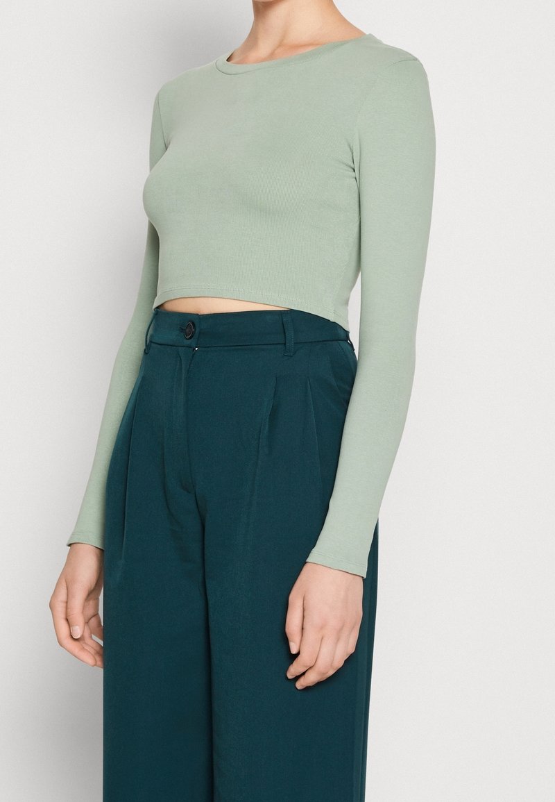Person iført en tætsiddende lysegrøn langærmet crop top og højtaljede mørk teal vide bukser, stående mod en ensfarvet baggrund.
