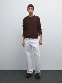 Maglione bordeaux scuro con colletto grigio, abbinato a pantaloni bianchi a gamba dritta e scarpe in suede marroni, su uno sfondo minimalista.