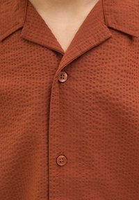 Roestkleurig shirt met korte mouwen van een gestructureerde stof. Voorzien van een geknipt kraagje en twee knopen aan de voorkant.