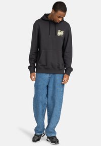 Hoodie noir avec poche avant, capuche à cordons et graphique jaune. Associé à un jean bleu loose et des chaussures de sport noires.