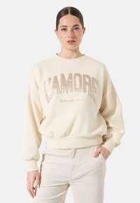 Sudadera color crema con gran texto "L'AMORE" en beige, puños acanalados y un corte relajado. Presenta una tela suave y un cuello redondo.