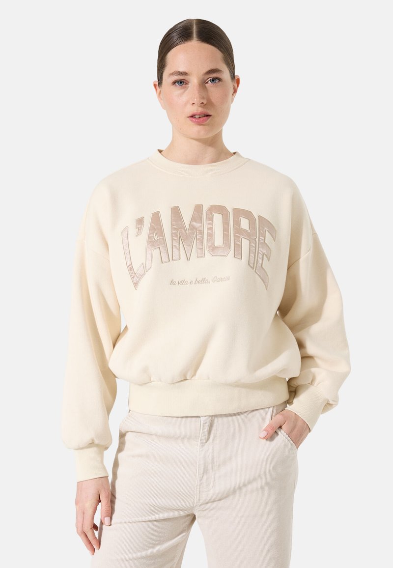 Sudadera color crema con gran texto "L'AMORE" en beige, puños acanalados y un corte relajado. Presenta una tela suave y un cuello redondo.