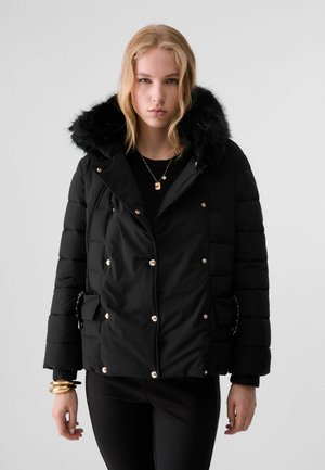 Junge Frau mit blondem Haar, die eine schwarze Steppjacke mit pelzgefütterter Kapuze und goldenen Knopfdetails trägt, steht vor einem schlichten Hintergrund.
