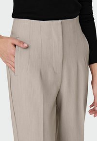 Pantalon beige ajusté avec plis devant, poches latérales et texture lisse, présenté avec une main placée dans une poche.
