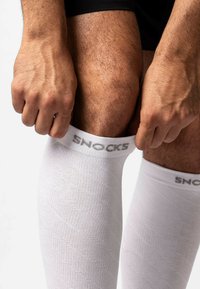 Calze a compressione bianche con motivo texturizzato. Il nome del marchio "SNOCKS" è visibile vicino alla parte superiore, indossate su gambe di una persona.