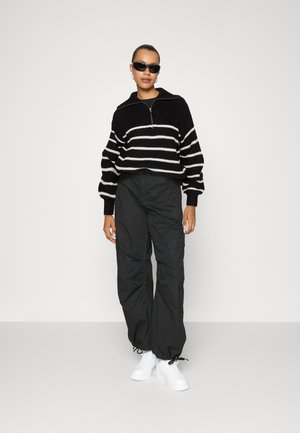 Pull en tricot à rayures noires et blanches avec un col zippé, associé à un pantalon cargo noir et des baskets blanches. Texture lisse dans l'ensemble.