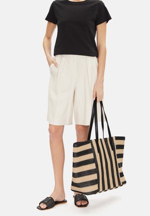 Donna che indossa una t-shirt nera, pantaloncini beige fino al ginocchio, sandali neri a ciabatta e tiene in mano una grande borsa a tracolla a righe nere e beige.