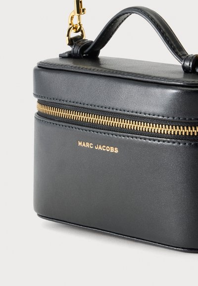 Μαύρη δερμάτινη τσάντα Marc Jacobs με χρυσό φερμουάρ, χρυσά λογότυπα, επάνω χερούλι και χρυσά μεταλλικά στοιχεία σε απλό ανοιχτόχρωμο φόντο.