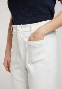 Pantalon blanc avec une texture lisse, doté de grandes poches plaquées et d'un bouton, associé à un haut foncé et texturé.