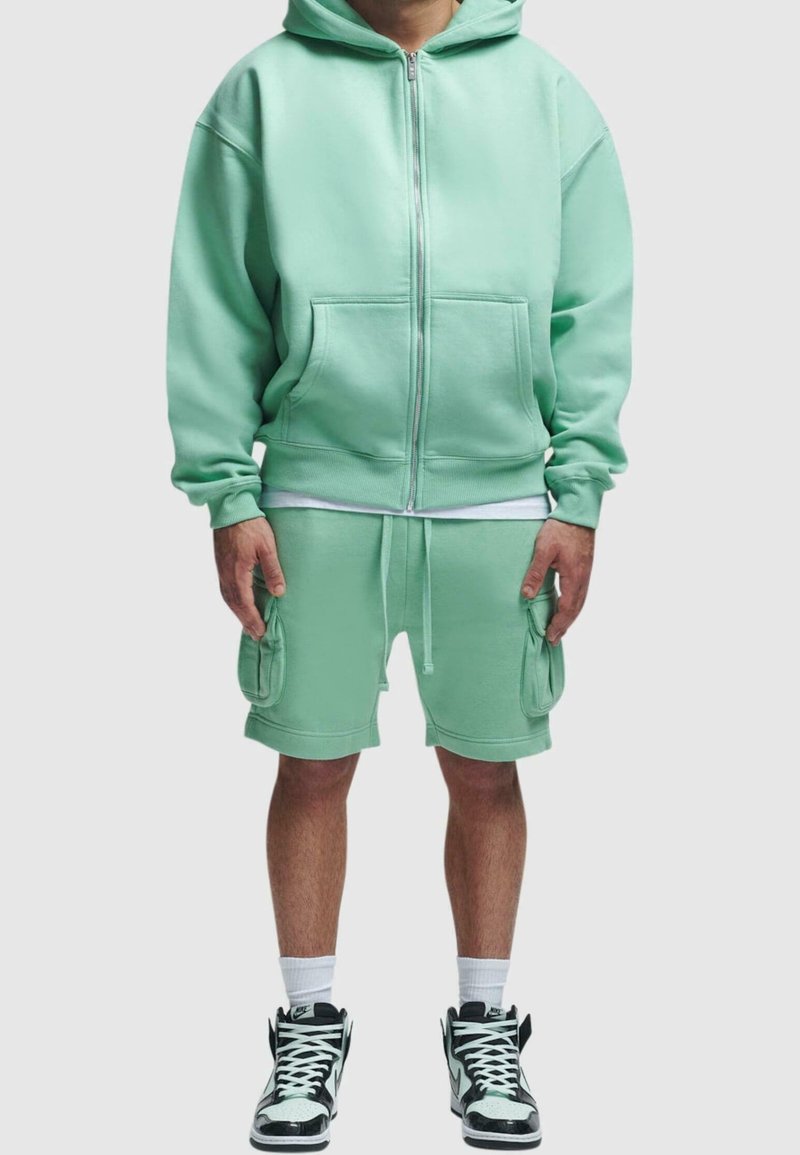 Homme portant un sweat à capuche zippé vert menthe et un short cargo assorti, avec des chaussettes blanches et des baskets montantes noir et blanc.