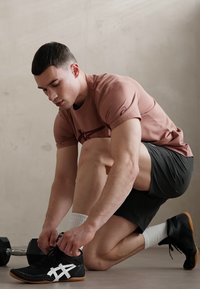 Atletische man op zijn knieën die zwarte sportschoenen met witte accenten vastbindt. Hij draagt een lichtroze T-shirt en zwarte shorts in een sportschoolomgeving.