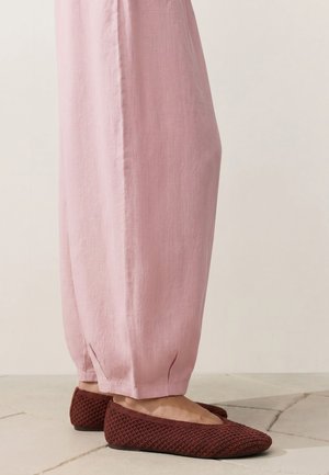 Person trägt lockere rosa Hose und kastanienbraune, strukturierte Ballettschuhe und steht auf einem hell gekachelten Boden.