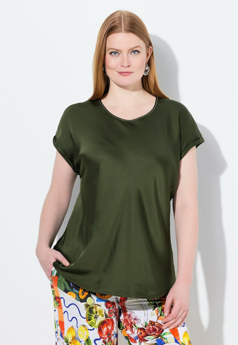 Olivgrübluse mit kurzen Ärmeln, runder Ausschnitt und glatter Textur; kombiniert mit bunten, gemusterten Hosen mit Obstmotiven.