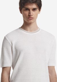 Vit kortärmad stickad tröja med ett texturerat mönster och en ribbad rund halsringning, med lätt uppvikta ärmar.