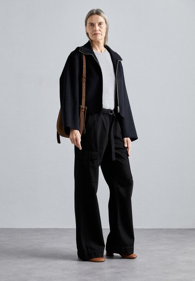 Black cape jacket with a silver zip, grey t-shirt, black wide-leg trousers, and brown handbag. Model stands on a gray background.