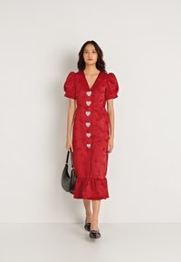 Κόκκινο φόρεμα με φουσκωτά μανίκια, κουμπιά σε σχήμα καρδιάς και ρέουσα τελείωμα. Φτιαγμένο από υφασμάτινο υλικό, συνδυασμένο με μαύρη τσάντα χειρός και φλατ παπούτσια.