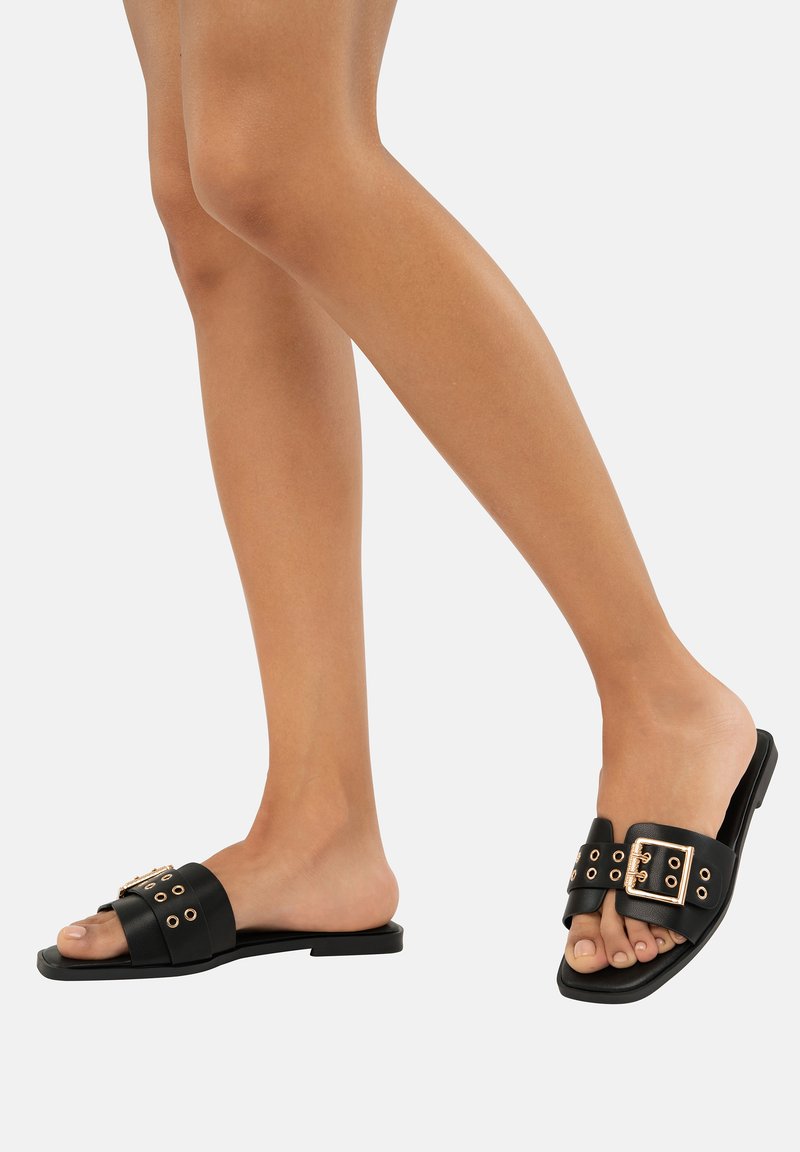 Infradito neri con una fascia ampia decorata con hardware dorato e occhielli, suola piatta e design open toe. Realizzati in materiale sintetico liscio.