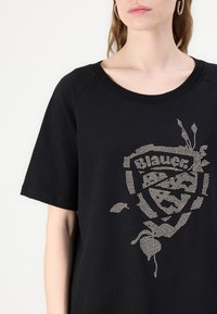 Zwarte t-shirt met korte mouwen, voorzien van een textuurdesign van rhinestones die een logo en bloemen elementen op de voorkant vormen.